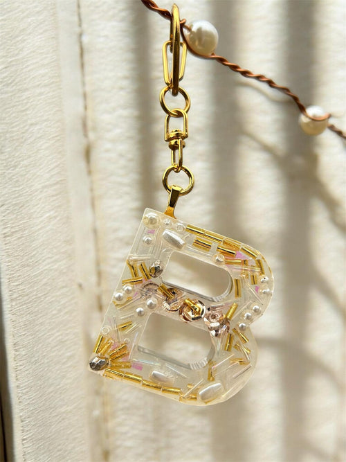 Clear and gold beaded decorative keychain letter "B" with pearls on a textured beige background