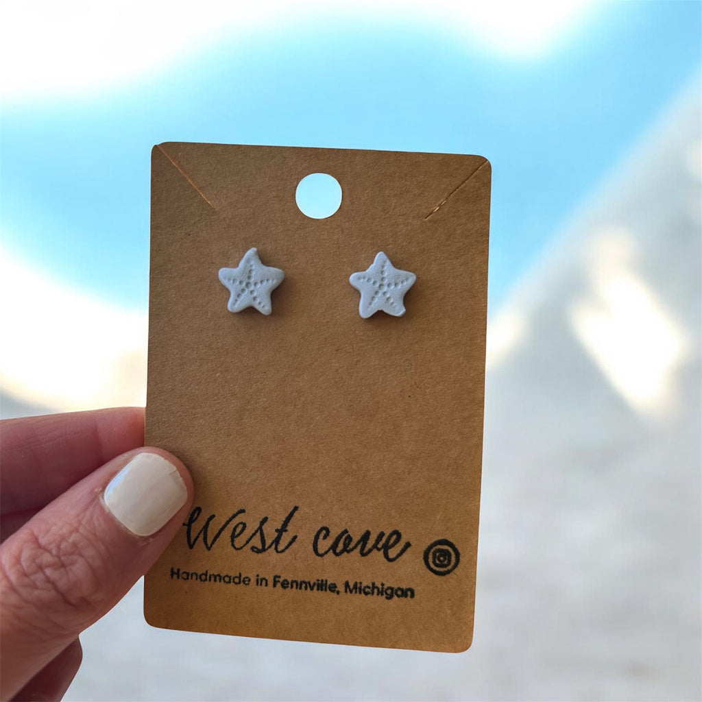 Pair of white polymer clay starfish stud earrings on a brown display card, coastal beach jewelry.